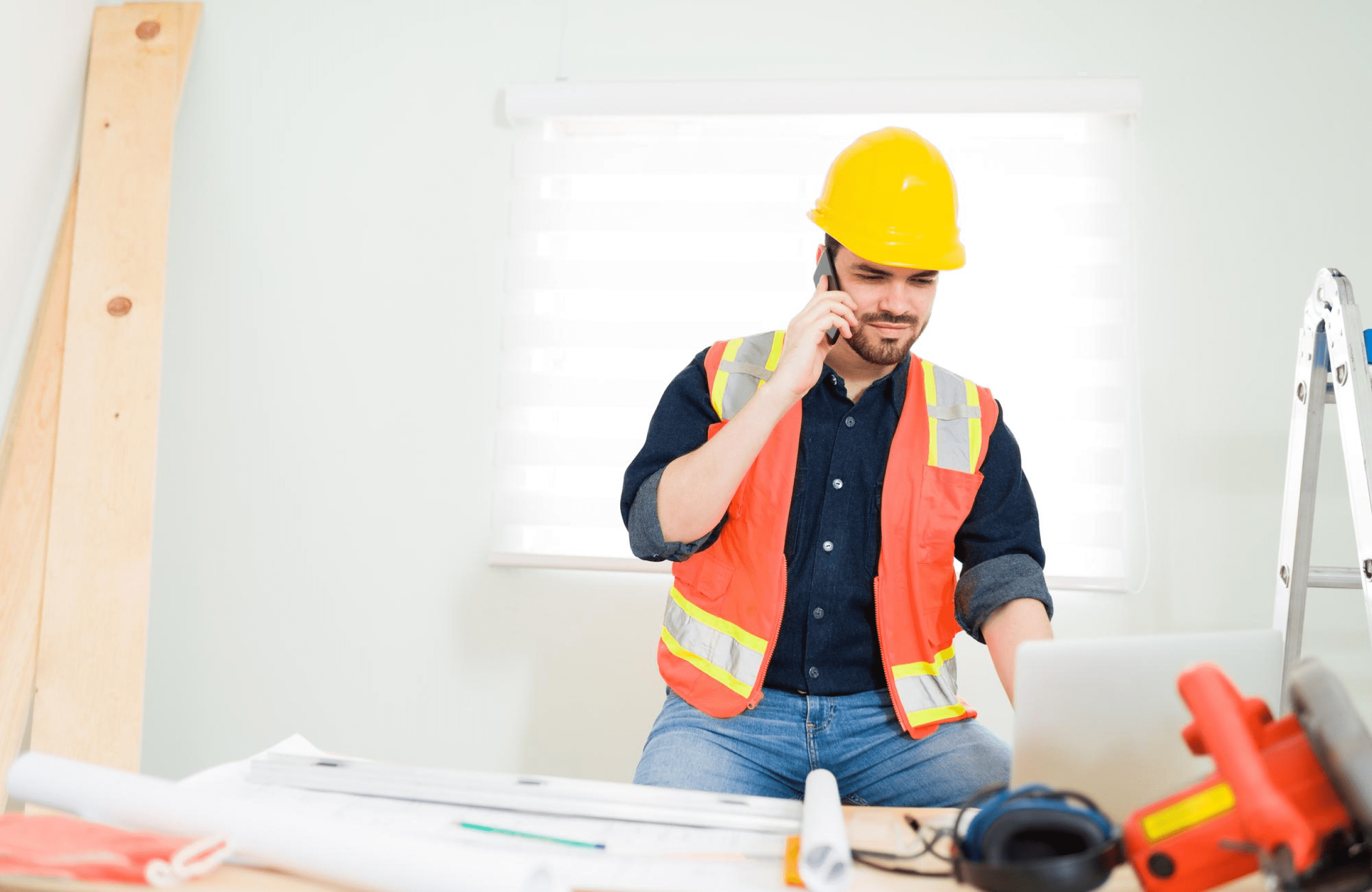 Stone Haven Developments A construction worker wearing a yellow hard hat and orange safety vest talks on the phone while using a laptop at a desk with blueprints and tools. Ontario