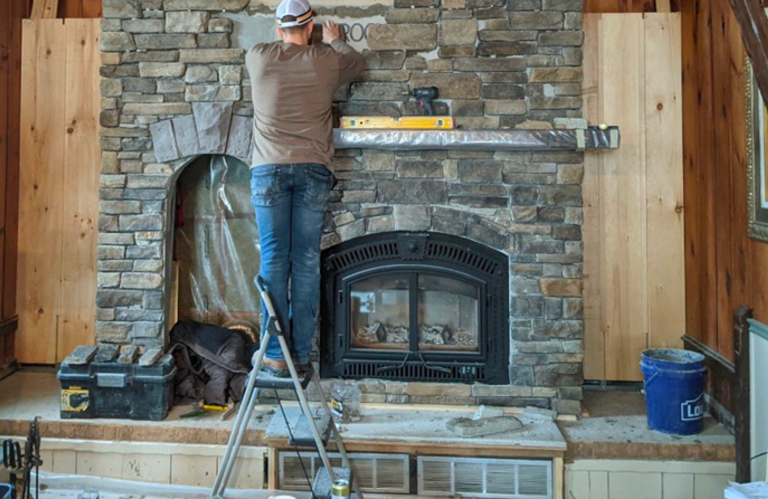 Stone Haven Developments A person stands on a ladder installing or working on a stone fireplace; tools, a toolbox, and a blue bucket are nearby in a partially finished room. Ontario